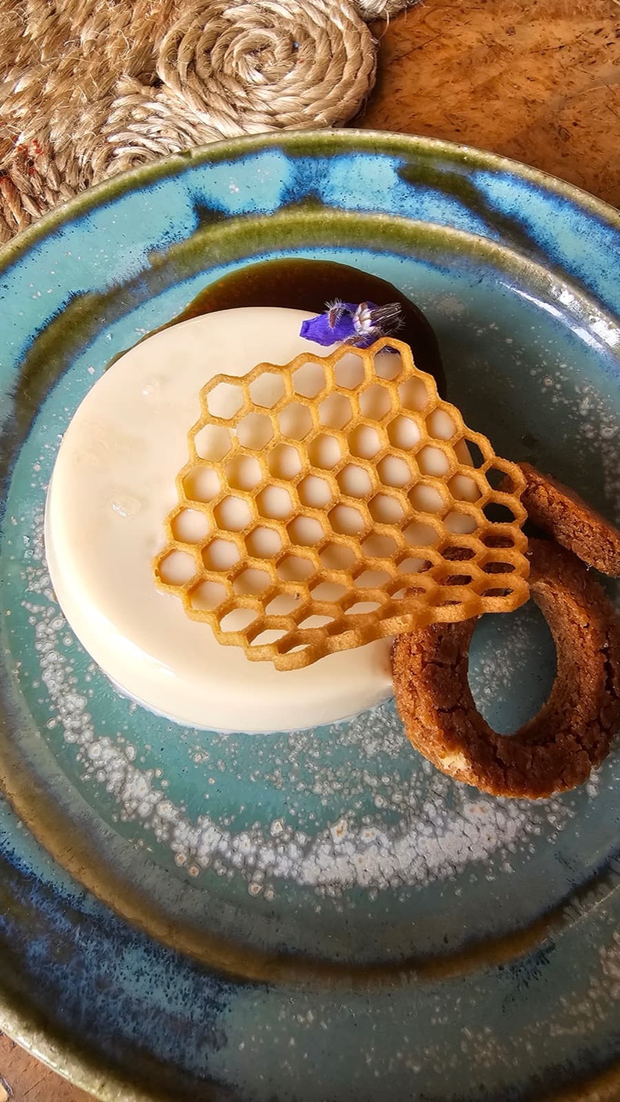 Set cream dessert on a blue-glazed plate, topped with a honeycomb-patterned tuile and a purple edible flower, with a curved biscuit alongside